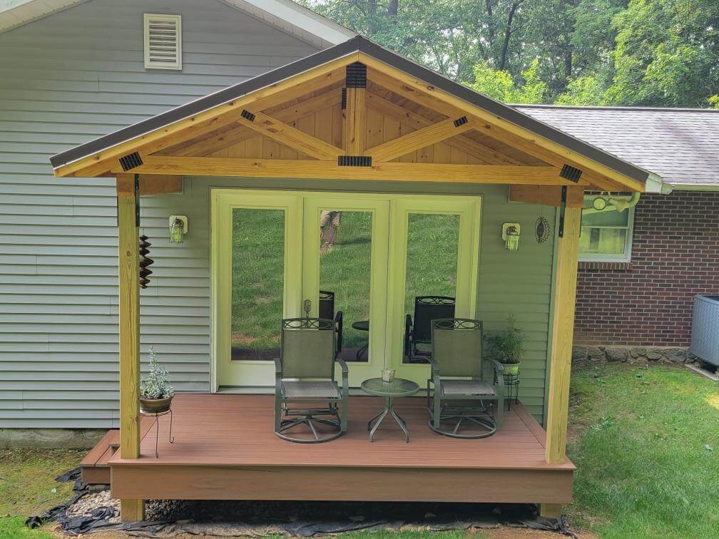 A small deck with two chairs and a coffee table, featuring an a-frame overhang attached to the back of a house with two steps going off the side into the yard.